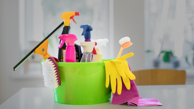 Secchio verde con guanti, detergenti e strumenti per le pulizie in condominio, posizionato su un tavolo in ambiente domestico luminoso.