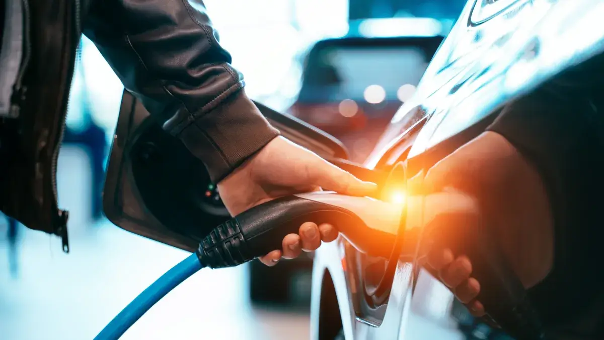Mano che collega un cavo di ricarica a un'auto elettrica, in un contesto urbano moderno, simbolo della ricarica elettrica in condominio.